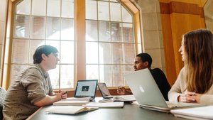 Three people sit at a table with laptops and notebooks, engaged in conversation near a large window.