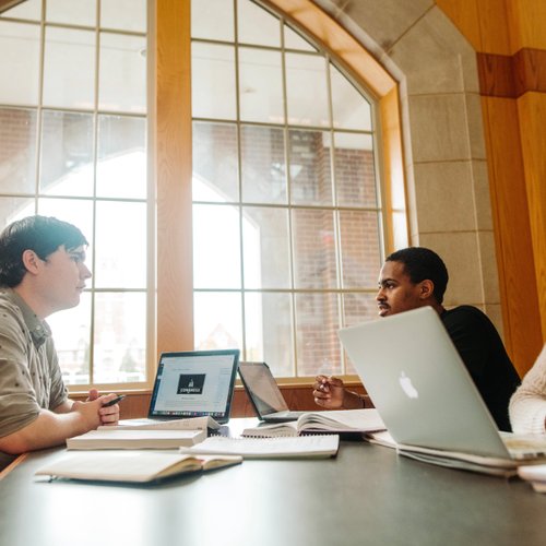 Three people sit at a table with laptops and notebooks, engaged in conversation near a large window.