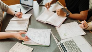 Three people study together at a table with open notebooks, textbooks, laptops, and pens.