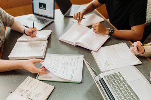 Three people study together at a table with open notebooks, textbooks, laptops, and pens.