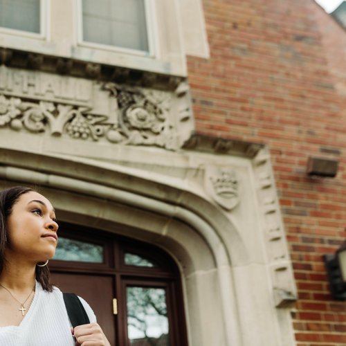 A young woman wearing a white sweater and cross necklace stands outside a brick building with ornate stonework above the