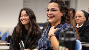 A smiling woman with glasses holds a small trophy while sitting next to a smiling woman in a classroom.
