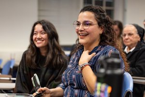 A smiling woman with glasses holds a small trophy while sitting next to a smiling woman in a classroom.