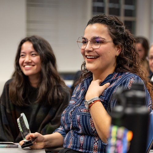 A smiling woman with glasses holds a small trophy while sitting next to a smiling woman in a classroom.