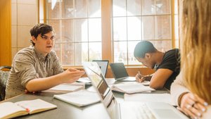 Three students study together at a table with laptops and notebooks in front of large windows.