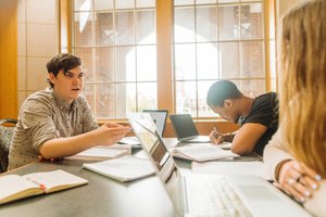 Three students study together at a table with laptops and notebooks in front of large windows.