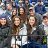 A group of smiling people dressed in jackets and hats sit closely together on bleachers at an outdoor event.