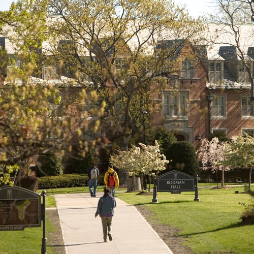 Three people walk along a paved path toward a large brick building partially obscured by trees and flowering shrubs.