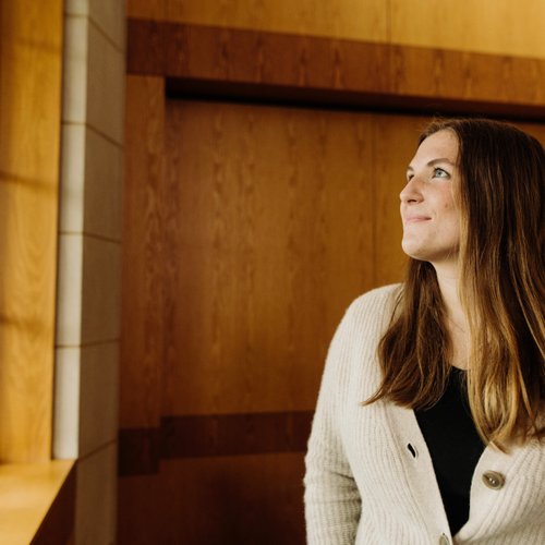 A woman with long brown hair wearing a beige cardigan looks out a window in a wood-paneled room.