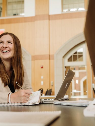 A woman with long brown hair smiles while writing in a notebook at a table with a laptop and open books.