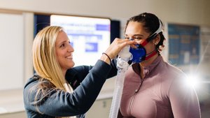A woman adjusts a blue breathing mask on another woman who is wearing a mauve zip-up jacket.