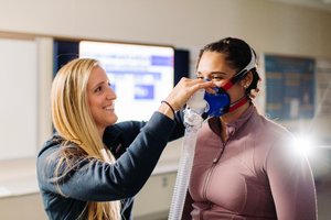 A woman adjusts a blue breathing mask on another woman who is wearing a mauve zip-up jacket.