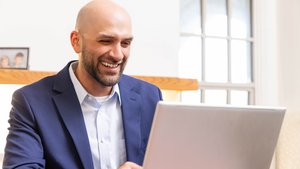 A smiling bald man in a blue suit jacket looks at a laptop screen in a bright room with a window behind him.