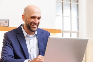 A smiling bald man in a blue suit jacket looks at a laptop screen in a bright room with a window behind him.