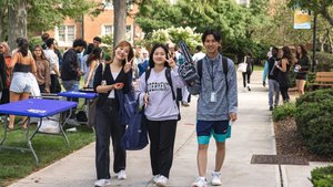 Three young adults walk on a campus path smiling and making peace signs while holding bags and a foam finger.
