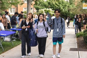 Three young adults walk on a campus path smiling and making peace signs while holding bags and a foam finger.