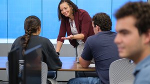 A woman leans on a table and gestures while talking to two seated people in front of a large blue screen.