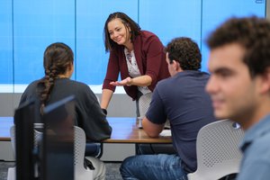 A woman leans on a table and gestures while talking to two seated people in front of a large blue screen.