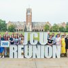 A group of people stands behind large lighted letters spelling JCU REUNION and a sign reading 2000 on a campus lawn with a