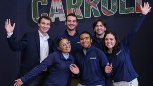 Six smiling young adults pose with raised hands in front of a dark blue wall with colorful "Greetings from John Carroll".