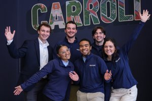 Six smiling young adults pose with raised hands in front of a dark blue wall with colorful "Greetings from John Carroll".