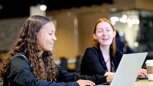 Two young women sit at a table working on laptops, one smiling and the other speaking.