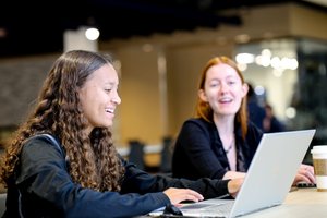 Two young women sit at a table working on laptops, one smiling and the other speaking.