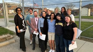 A group of eleven people stand closely together smiling in front of a chain-link fence with an American flag and buildings.