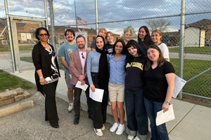 A group of eleven people stand closely together smiling in front of a chain-link fence with an American flag and buildings.