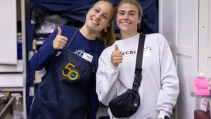 Two young women smile and give thumbs up while standing indoors near shelves and storage bins.