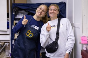Two young women smile and give thumbs up while standing indoors near shelves and storage bins.