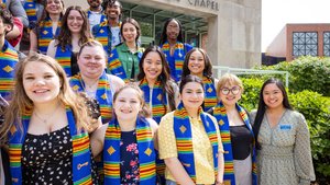 A diverse group of smiling students wearing blue and yellow stoles stand outside Saint Francis Chapel on a sunny day.