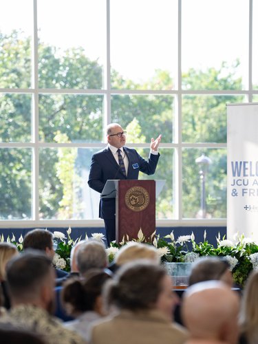 A man in a suit speaks at a podium in front of an audience with banners and flowers behind him.