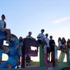 A group of nine people sit and stand on large colorful letters spelling Belize against a clear sky at sunset.