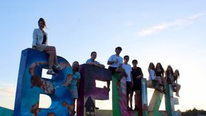A group of nine people sit and stand on large colorful letters spelling Belize against a clear sky at sunset.