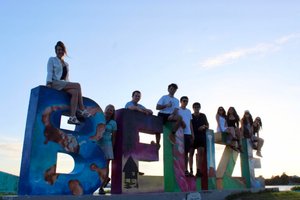 A group of nine people sit and stand on large colorful letters spelling Belize against a clear sky at sunset.