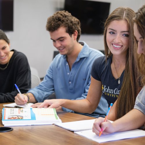 Four young adults sit around a table smiling and writing in notebooks during a group study session.