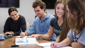 Four students sit at a table smiling and writing in notebooks during a group study session.