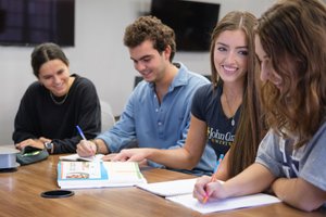 Four students sit at a table smiling and writing in notebooks during a group study session.