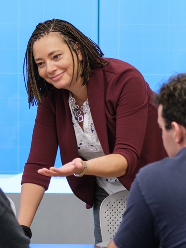 A woman in a maroon jacket leans forward speaking to two seated people while another person is blurred in the foreground.