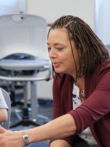 A woman in a maroon jacket points at a notebook while two young women watch and smile in a classroom.