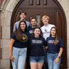 Six young adults smile standing closely together in front of a large wooden arched door set in a brick and stone wall.