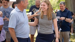 A man in a light blue shirt smiles and touches the shoulder of a smiling young woman in a gray shirt and navy skirt outdoors.