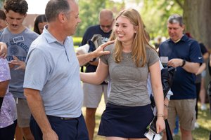 A man in a light blue shirt smiles and touches the shoulder of a smiling young woman in a gray shirt and navy skirt outdoors.