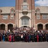 A large group of people, including many in academic robes, pose for a photo in front of a tall brick building with arched.
