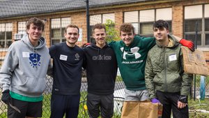 Five young men stand close together smiling in front of a garden fence, two holding paper bags and gardening tools.