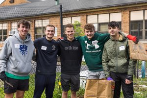 Five young men stand close together smiling in front of a garden fence, two holding paper bags and gardening tools.