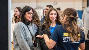 Three women are engaged in conversation indoors, with one woman gesturing while the others listen and smile.