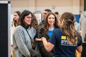 Three women are engaged in conversation indoors, with one woman gesturing while the others listen and smile.