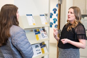 A woman in a black top gestures while talking to another woman in a gray jacket near a rack of brochures.